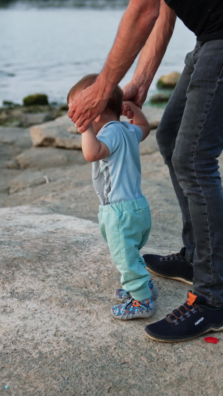 Man wearing jeans leading his little son studying to walk. Beautiful baby boy in blue clothes stepping by stones. Bank of the river setting. Vertical video
