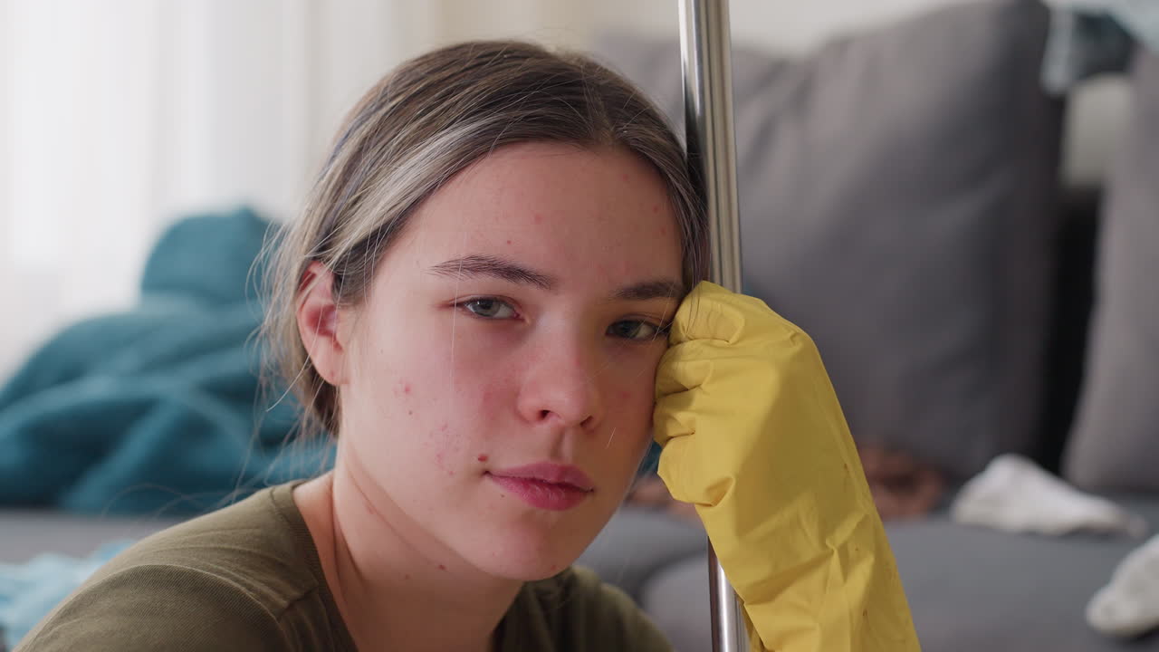 Young woman wearing yellow gloves resting head against mop stick seated in messy living room with sofa and scattered clothes in background looking forward with calm expression