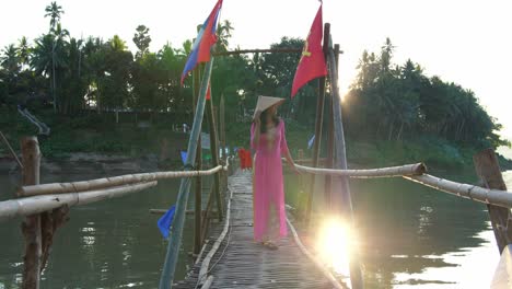 woman in pink ao dai on a bamboo bridge over a river at sunrise/sunset