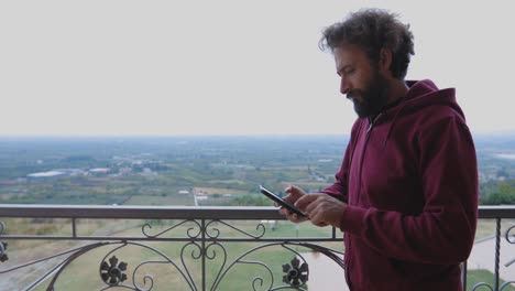 hombre revisando su teléfono inteligente mientras está parado en el balcón con una vista impresionante