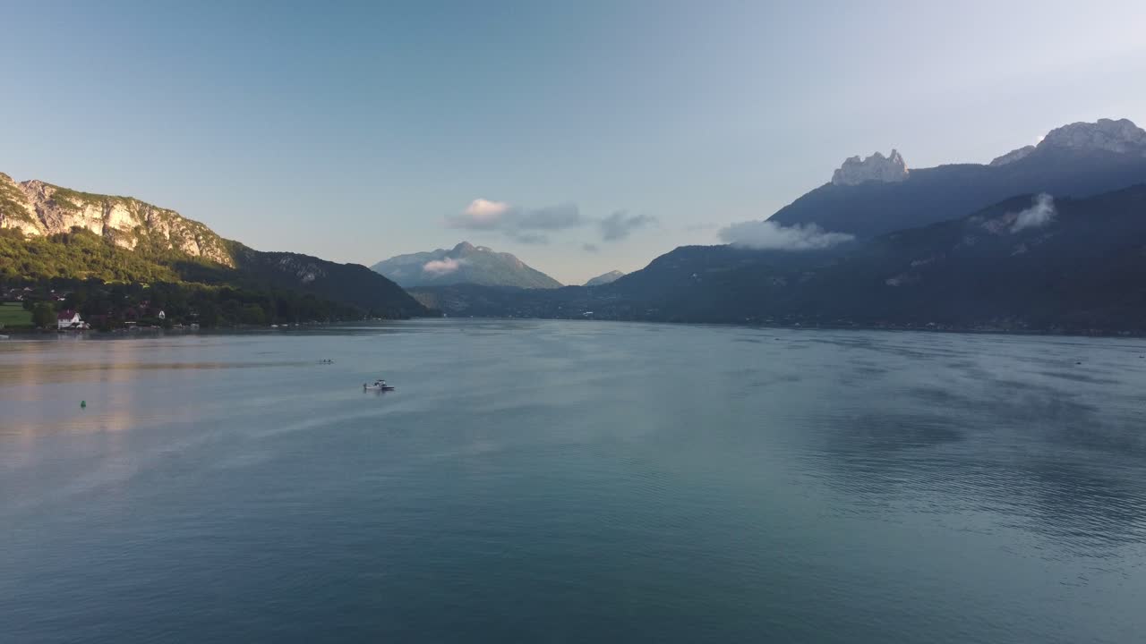 Rotational drone footage of Lake Annecy just after Sunrise. The sun has just appeared over the mountain tops and the odd boat is already on the water. Footage moves to the left. Clouds on the hills
