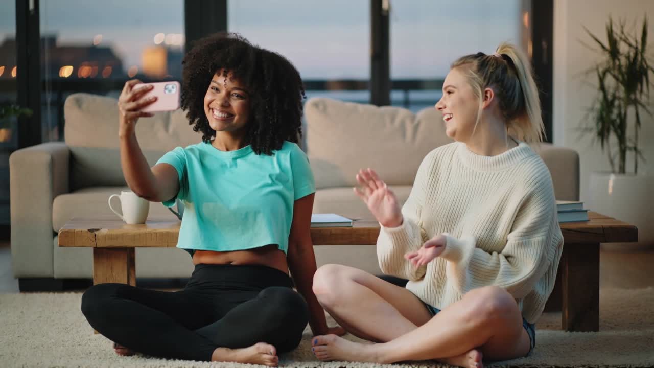 Holding smartphone, two women in teal and cream waving, laughing, filming selfie in living room