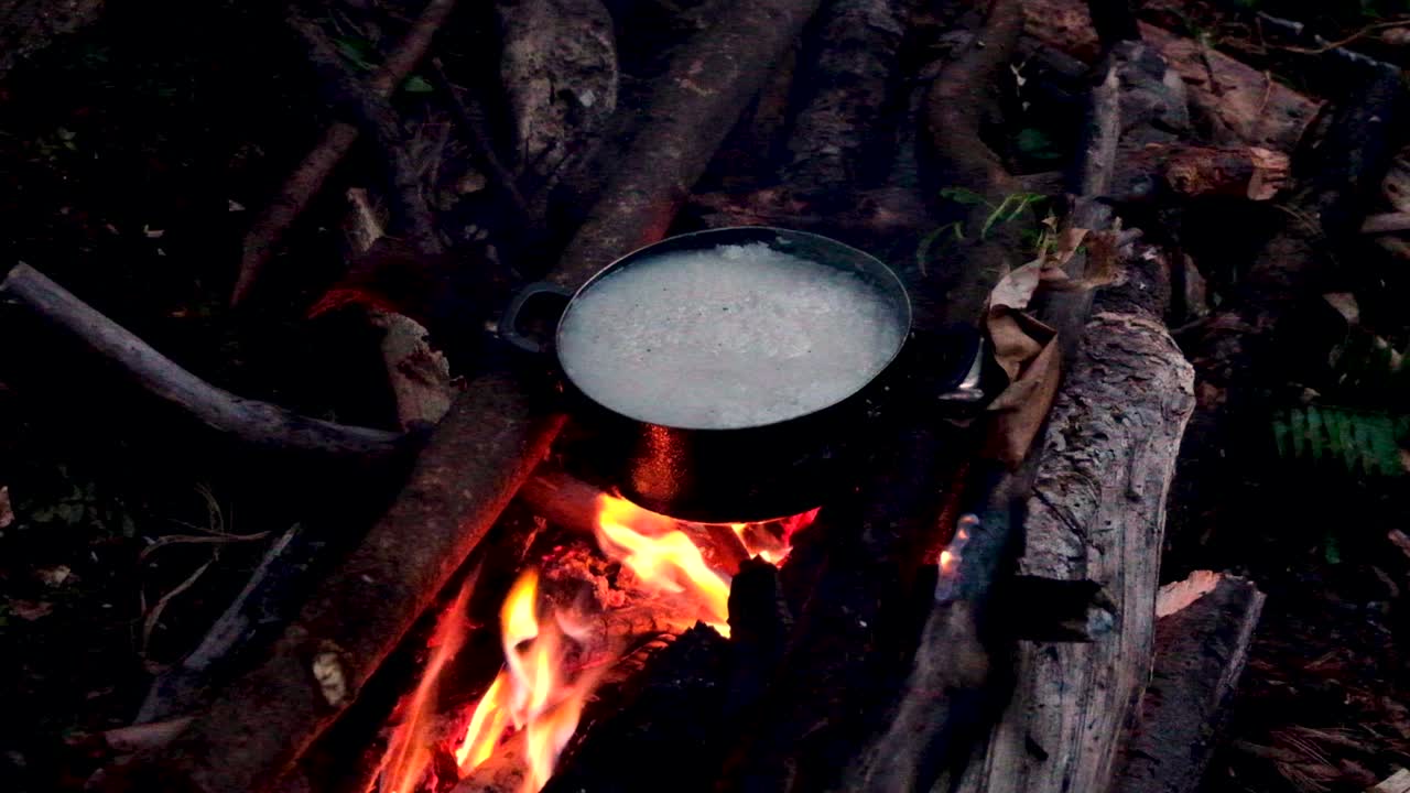 Uma panela de arroz branco cozinhando em fogo aberto ao ar livre na floresta tropical com toras e chamas, durante uma caminhada noturna em uma ilha tropical remota.