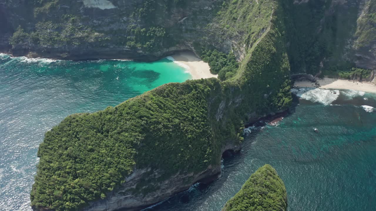Aerial drone shot in the iconic Kelingking Beach, the 'T-Rex' shaped headland, and the towering limestone cliffs of Nusa Penida. Turquoise Indian Ocean crashes against the rocks and white-sand beach