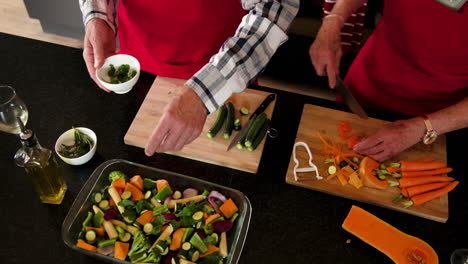Senior couple preparing fresh vegetables together in kitchen, wearing red aprons, at home