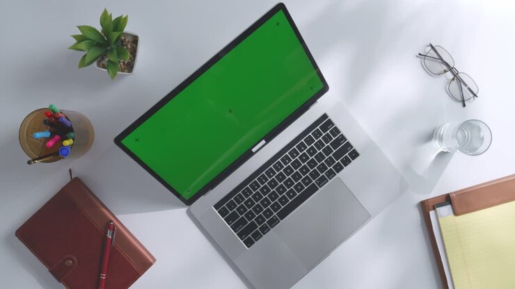 Draufsicht auf einen Laptop-Computer mit Mockup-Greenscreen-Display auf einem Holzschreibtisch, daneben ein Notizbuch mit Stiften, Brille und einem Glas Wasser. Langsames Herauszoomen, Nahaufnahme.