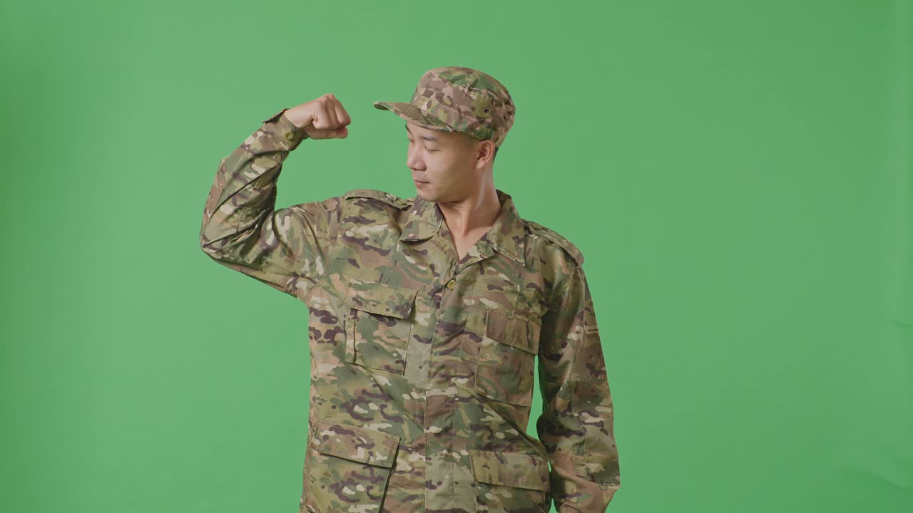 Asian Man Soldier Smiling And Flexing His Bicep While Standing In The Green Screen Background Studio