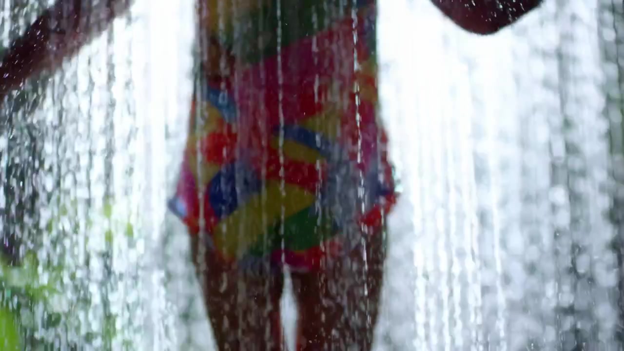 Cena em movimento vista por trás mostrando uma silhueta em pé atrás de uma densa queda d'água, com jatos translúcidos revelando quadris, pernas, braços e roupa de banho colorida e estampada com gotas brilhantes iluminadas pela borda e refrescante.