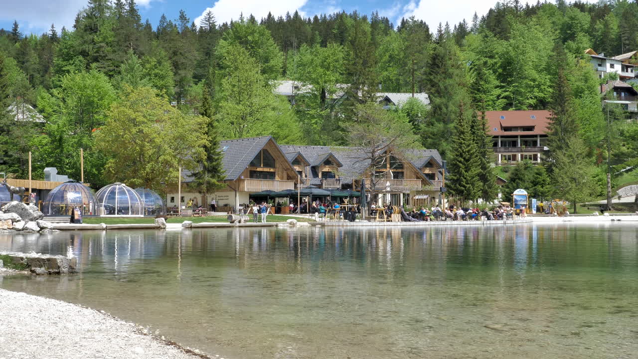 Lake Jasna in Slovenia with beautiful wooden houses and restaurant near water. Green and blue background with trees and clouds. 4k 50 fps shot
