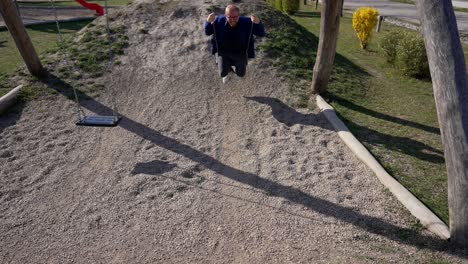 Adult-Man-Smiles,-Enjoys-Swings-in-Park,-Slow-Motion-on-Sunny-Day