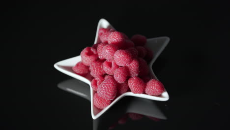 vibrant red raspberries in white star dish on black table with movement from left to right