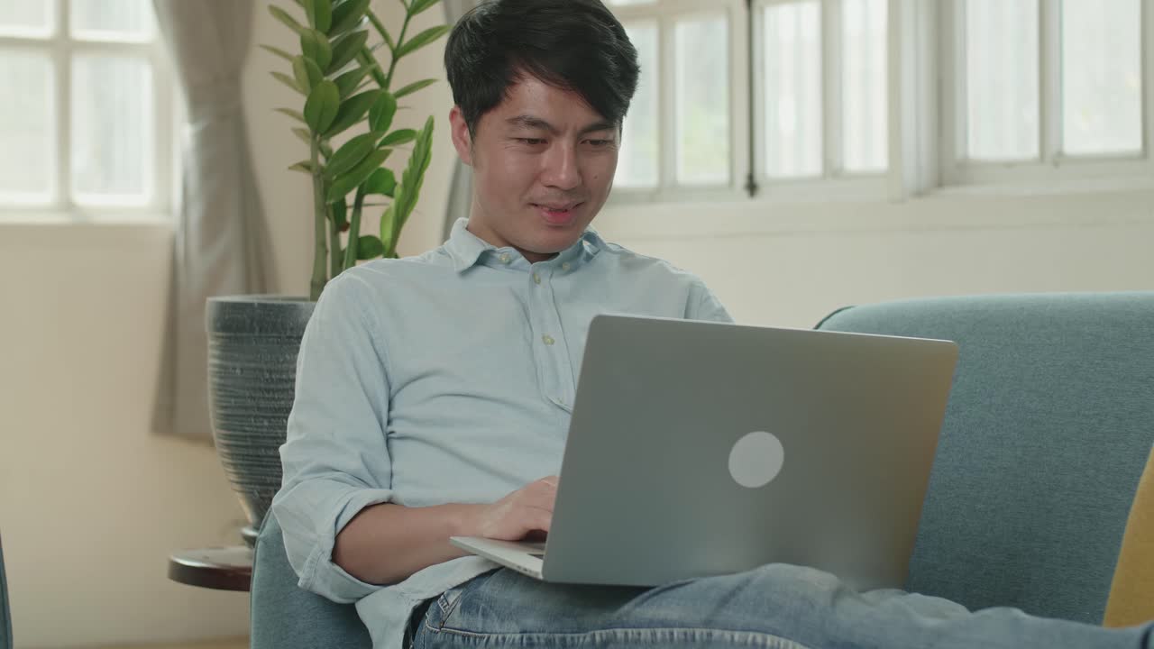 Asian Male Use Laptop Computer And Smiling In Living Room While Lying On Sofa, Video In 4K