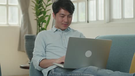 asian male use laptop computer and smiling in living room while lying on sofa, video in 4k