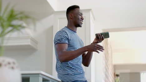 Feliz-Hombre-Afroamericano-Usando-Un-Teléfono-Inteligente-Y-Riéndose-En-Una-Cocina-Soleada,-Cámara-Lenta