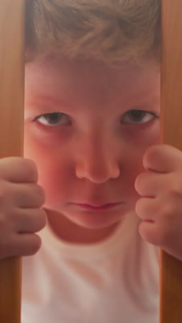 Serious little boy eats snack grabbing wooden bars of crib with hands. Adorable toddler chews in nursery looking in camera closeup slow motion