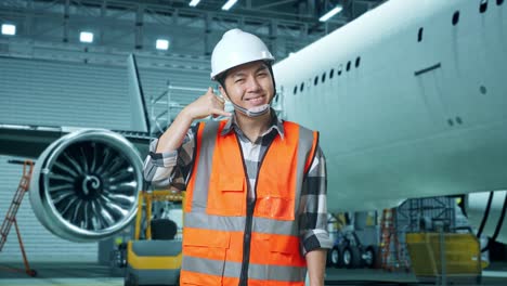 asian male engineer with safety helmet standing with aircraft in the hangar. smiling to camera and making call me gesture while aircraft maintenance