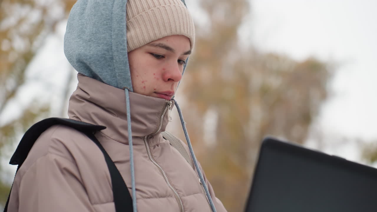 Light-skinned woman wearing hoodie and winter jacket stands outside on cold day, intently focused on laptop screen while working, with soft background of trees and neutral expression on her face