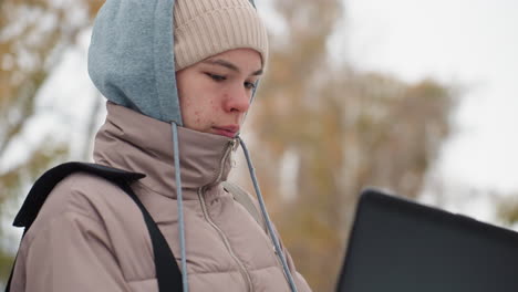 light-skinned woman wearing hoodie and winter jacket stands outside on cold day, intently focused on laptop screen while working, with soft background of trees and neutral expression on her face