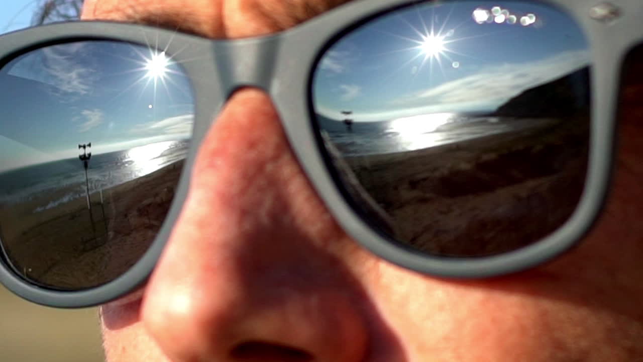 close-up shot of a person’s sunglasses reflecting a sunny beach and ocean waves. The reflection captures the beauty of the sea, sun, and sky in a unique and artistic way.
