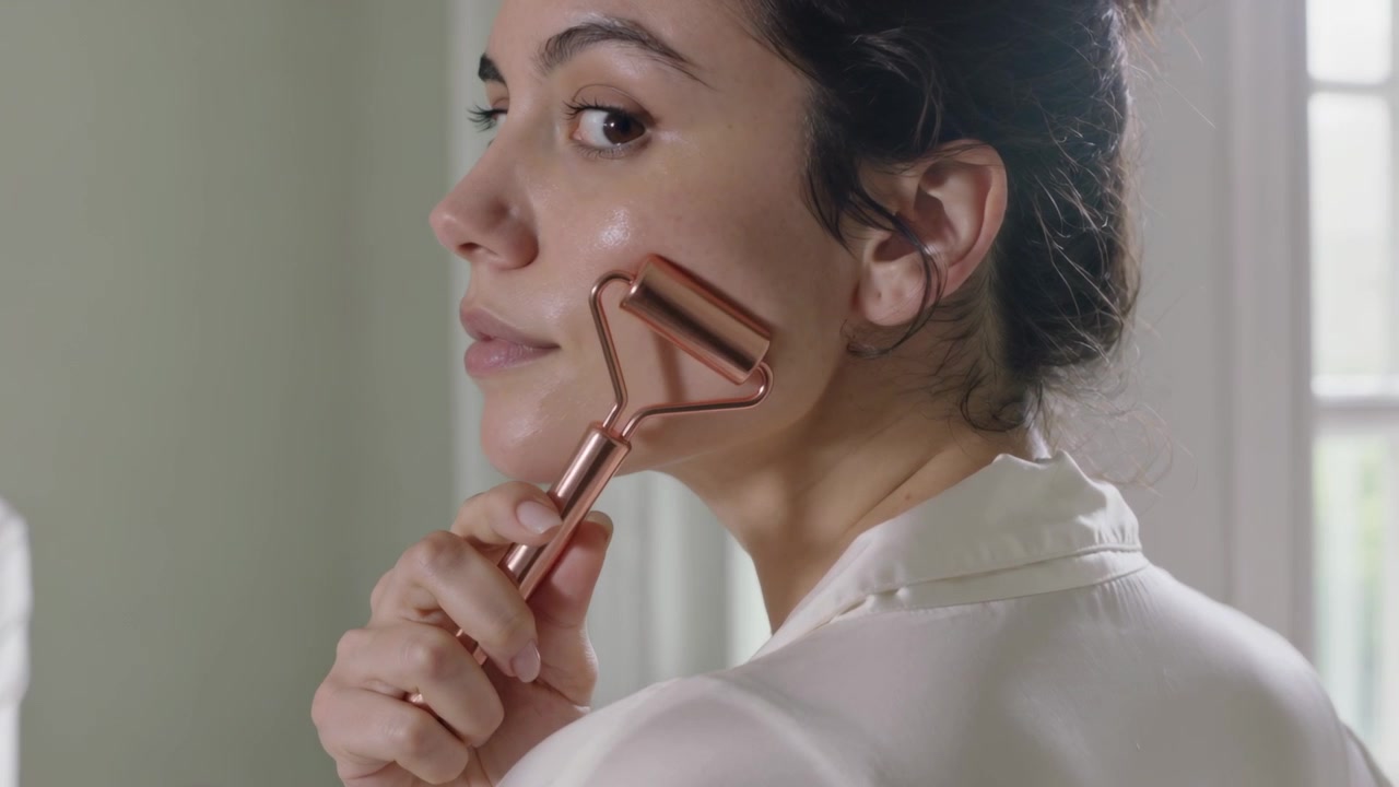 Una donna inizia a tenere un rullo color oro rosa, facendolo scorrere lungo la mascella davanti allo specchio di casa per la cura della pelle, in accappatoio di seta.