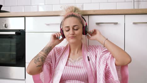 happy young woman with pink hair sitting on the floor on kitchen wearing pink pajamas and listening to music with headphones