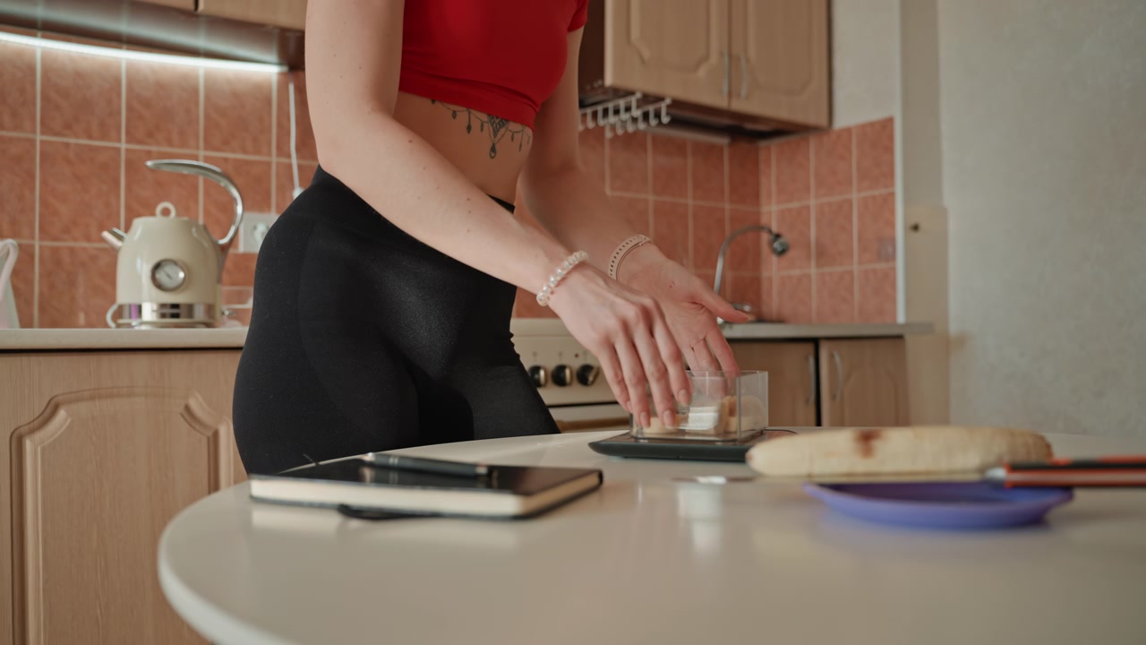 Primo piano delle mani sul bancone della cucina che preparano toast dopo l'allenamento, dita che spalmano burro con un coltello su una fetta, tagliere e piatto nelle vicinanze, telefono e taccuino sul tavolo, paraschizzi in piastrelle calde, tranquillo
