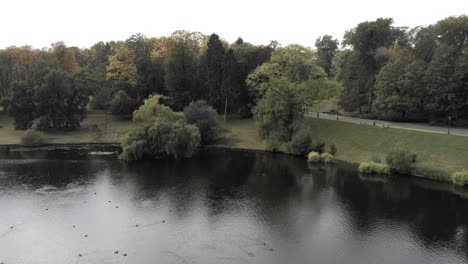 La-Lluvia-Comienza-A-Caer-En-Un-Lago-Del-Parque,-Los-Colores-Del-Otoño-Se-Reflejan-En-La-Superficie-Del-Agua,-La-Plataforma-Rodante-Aérea-Vuelve