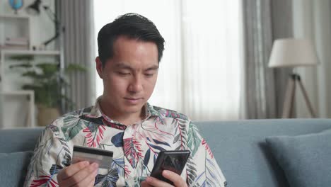 asian man sitting on couch in living room holding credit card and using smartphone shopping online