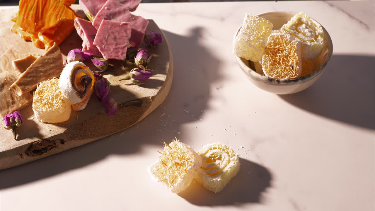 A hand selects a yellow swirled Turkish delight cube garnished with crispy kataifi dough from a group of sweets on a sunlit marble surface, with rosebuds, chocolate visible on a rustic wooden board