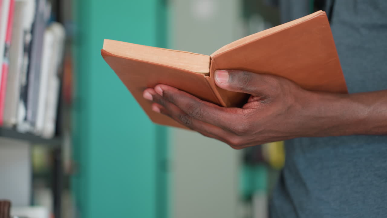 Close up of person adjusting and holding open old book in library, showing hands and worn cover with background of blurred colorful shelves