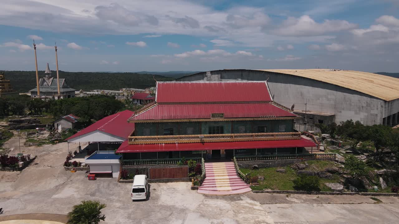 Traditional asian building with long red roofs at Bokor Hill Station with a golden statue and temples in Cambodia in the background. Drone panning shot