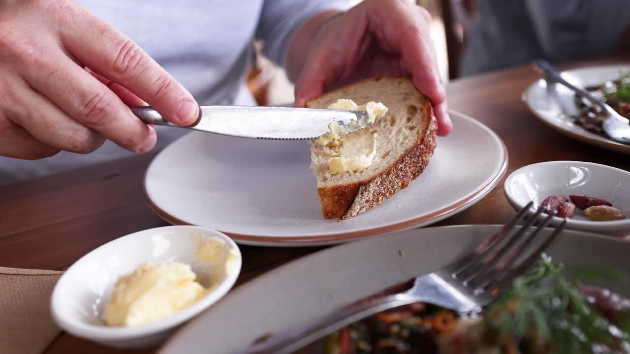 A person spreads butter on a slice of bread at a dining table, captured in natural lighting
