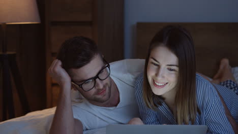 young smiling caucasian man and woman lying on the bed, talking and using a laptop at night