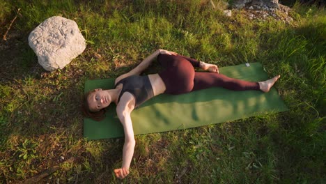 Woman-doing-supine-spinal-twist-yoga-pose-in-forest,-morning-sunrise-in-nature