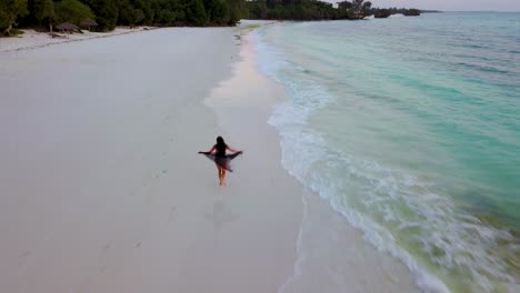 Aerial-drone-shot-of-a-woman-model-running-freely-along-a-white-sand-tropical-beach-in-a-flowing-dress-or-skirt