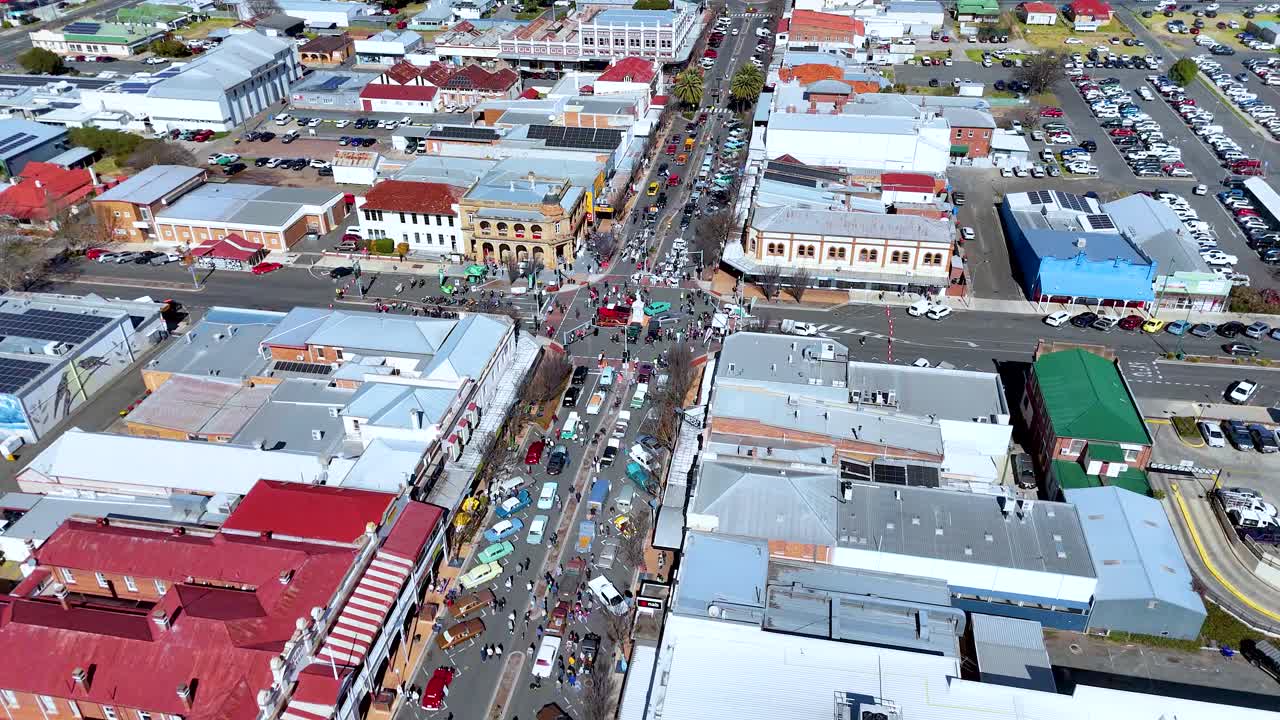 High-angle drone footage shows a vibrant town street lined with historic buildings, crowds, and market stalls under bright daylight, with smooth forward camera movement