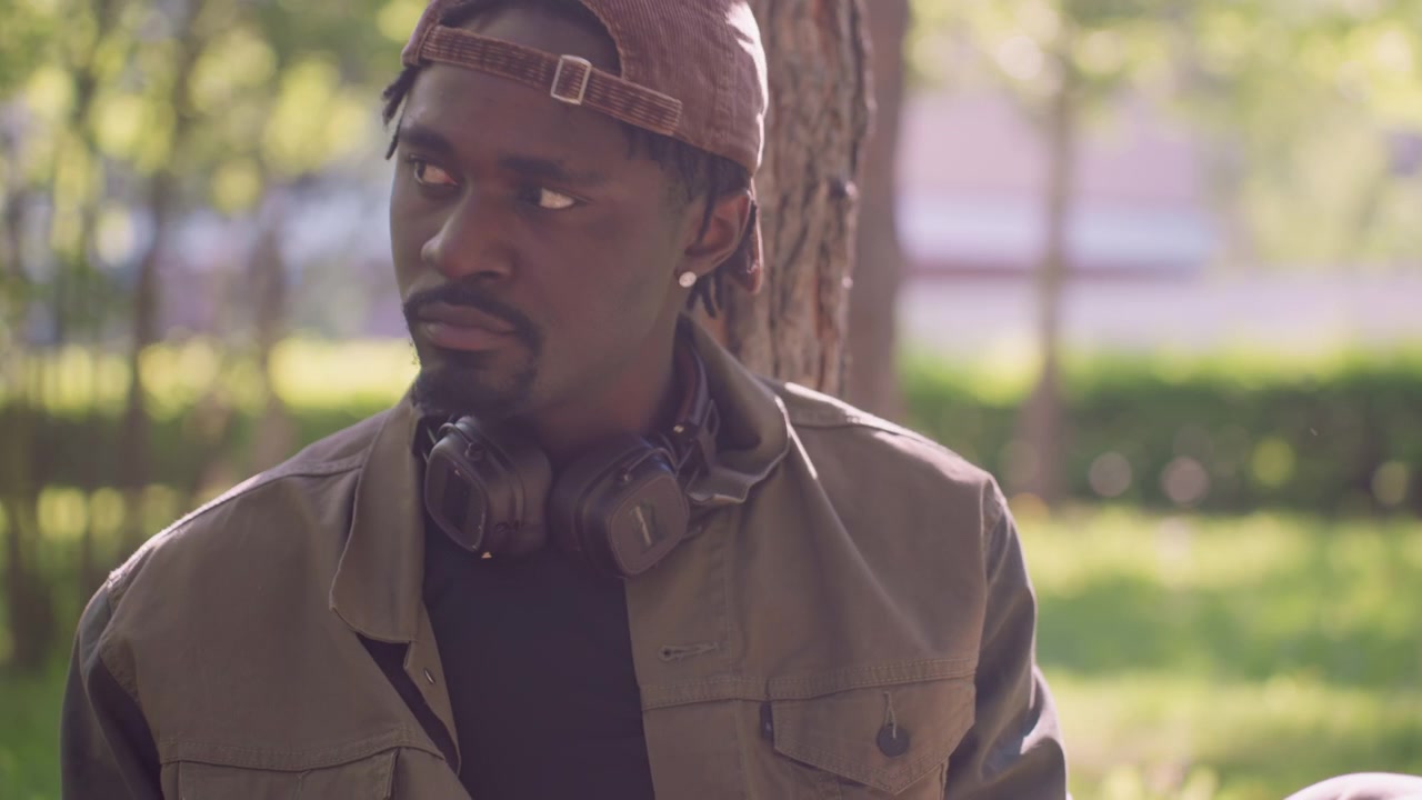 Primo piano di un uomo seduto all'aperto che legge un libro aperto in una luce naturale soffusa. L'uomo indossa cuffie al collo e siede sull'erba con alberi visibili dietro di lui. La scena è calma e focalizzata.