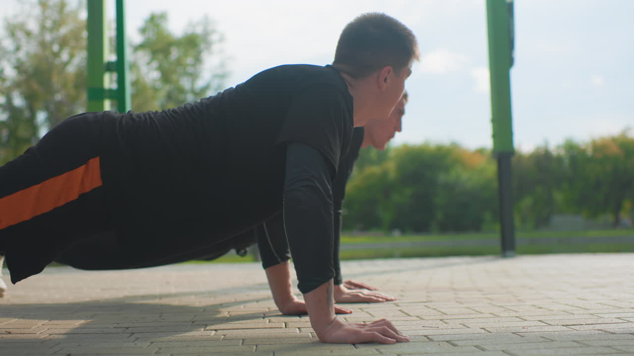 Man performing pushups outside in park, Closeup of man executing pushups in sunny autumn setting, Individual performing synchronized pushup exercises on pavement with natural park background