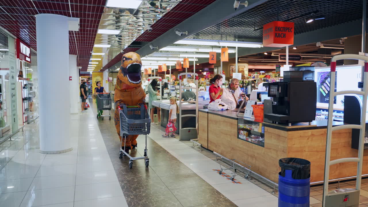 Tall orange dinosaur walking by supermarket pushing an empty cart. Funny T-Rex passing cash registers and leaves the cart.