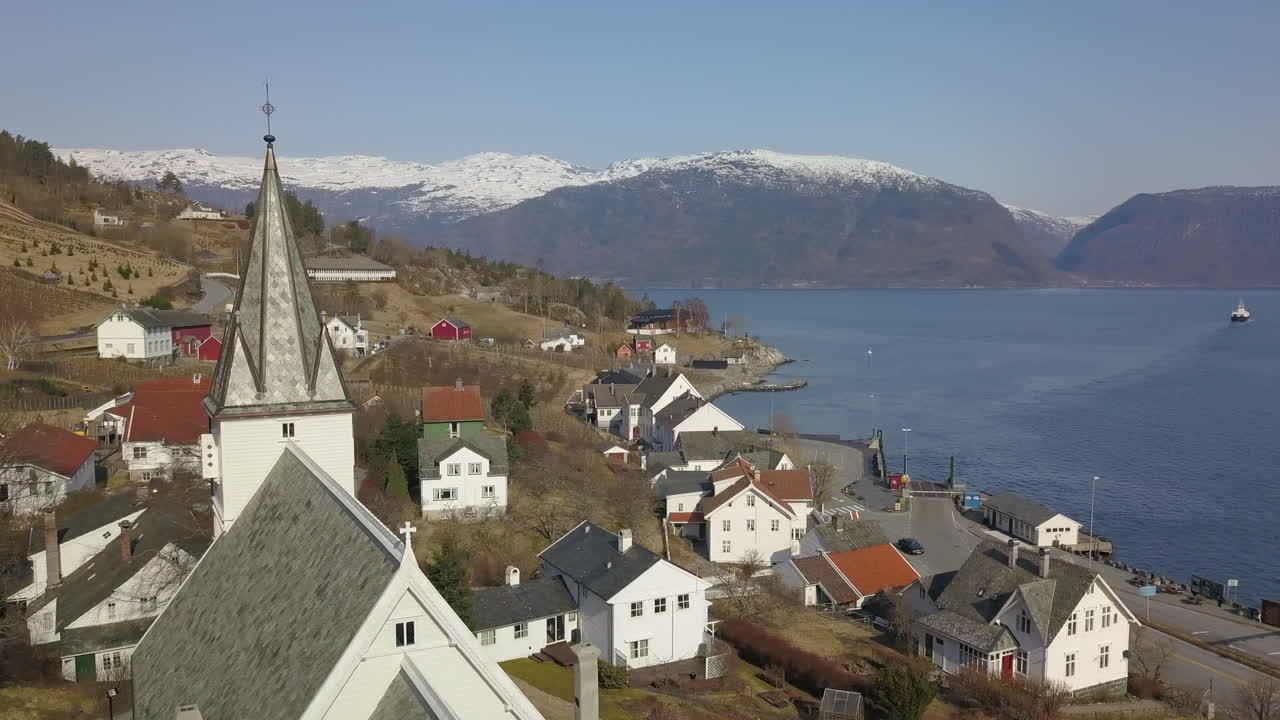 toma aérea orbitando alrededor de un campanario de iglesia en un pueblo noruego con montañas nevadas y un ferry cruzando un fiordo en el fondo