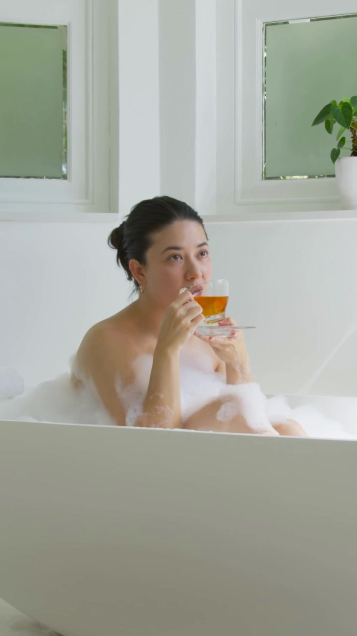 Vertical video: Settling in foam, woman holding cup, sipping tea to rest in tub with frosted panes