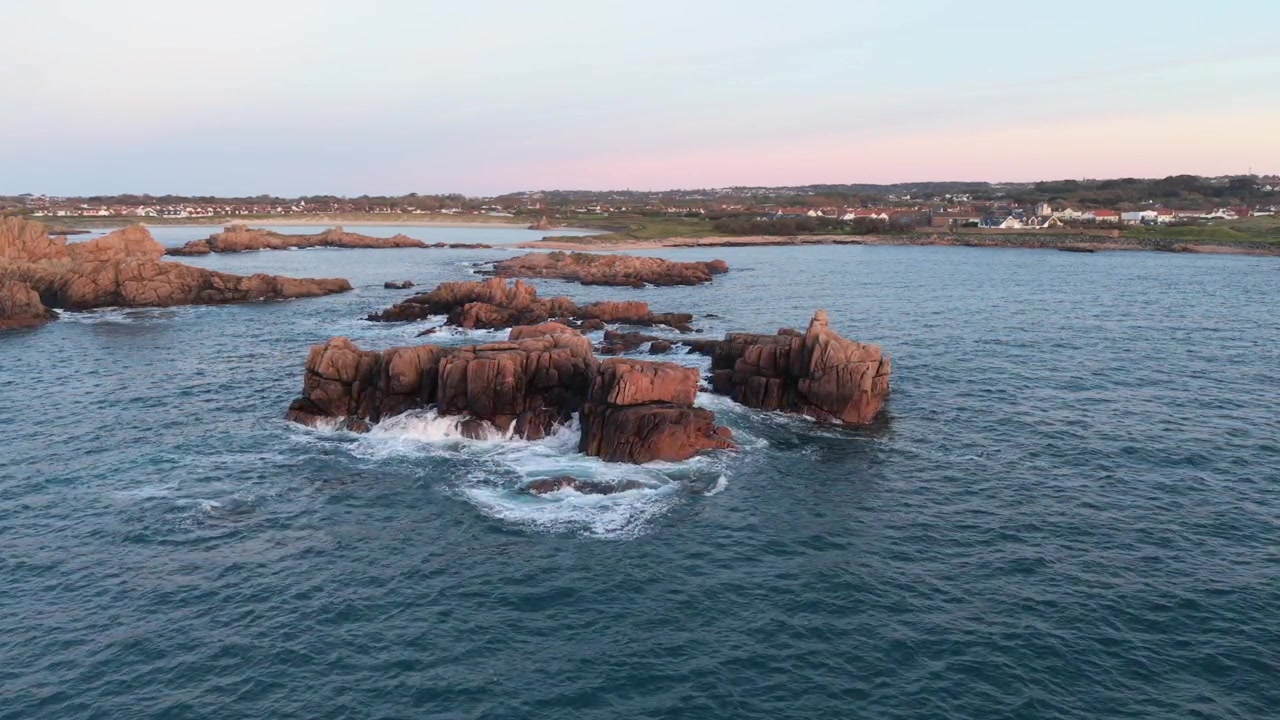 Guernsey, Isole del Canale, filmato ravvicinato con drone di onde spumeggianti che si infrangono su affioramenti rocciosi di granito circondati da mare calmo nel tardo pomeriggio, con il rosa che inizia a tingere l'orizzonte e viste della costa.