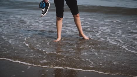 Niña-Descalza-Pateando-Agua-En-La-Playa-Sosteniendo-Zapatillas,-Cámara-Lenta