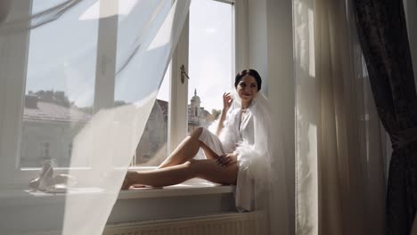 novia en vestido de boudoir sentada en el alféizar de la ventana preparaciones de la mañana de la boda mujer en vestidura de noche, velo