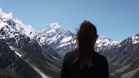 young caucasian woman looking around at sealy tarns viewpoint on a sunny summer day wih hooker lake, and snowy mountain peaks in background in mount cook national park, new zealand.