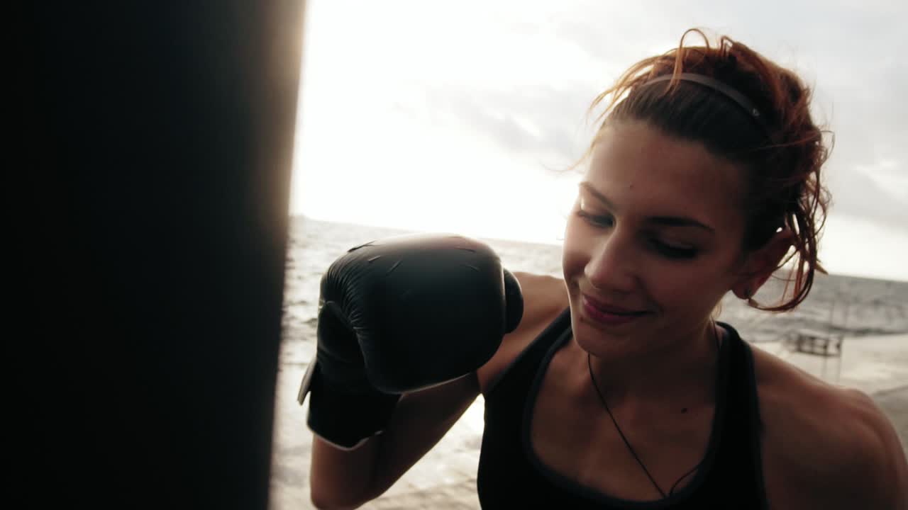 vista de cerca de una fuerte boxeadora atlética en guantes ejercitándose con una bolsa contra el hijo junto al mar. entrenamiento de boxeadora femenina
