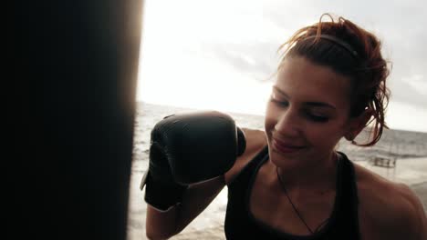 Close-Up-view-of-a-strong-athletic-female-boxer-in-gloves-exercising-with-a-bag-against-the-son-by-the-sea.-Female-boxer-training