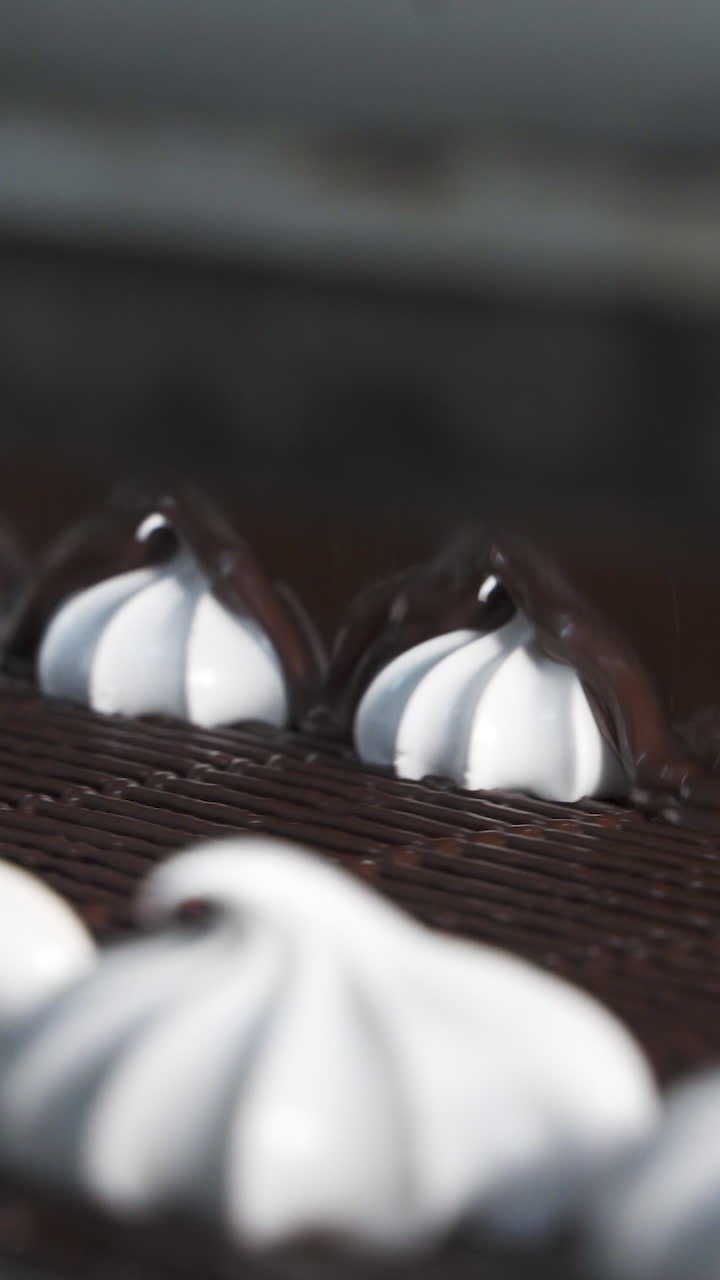 A white sweet marshmallow on a ribbon is covered with melted hot dark chocolate. Close-up. Conveyor at a confectionery factory. Vertical video