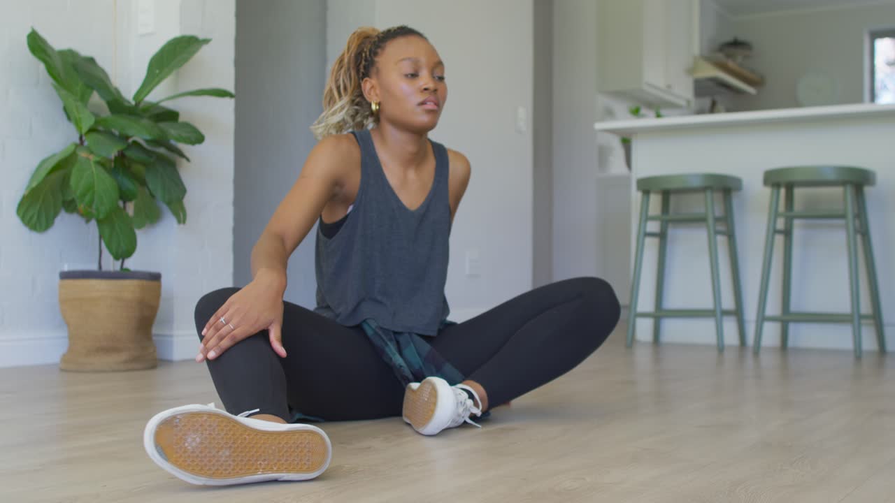 Donna afroamericana che esegue stretching da seduta nella zona giorno di casa, riscaldandosi con uno smartwatch