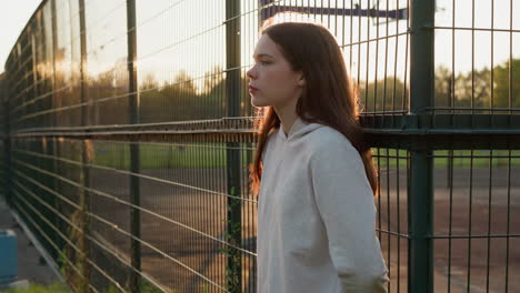 woman rests leaning on fence of stadium alone. young lady thinks about inner worries after jogging at back sunset. loneliness atmosphere in evening